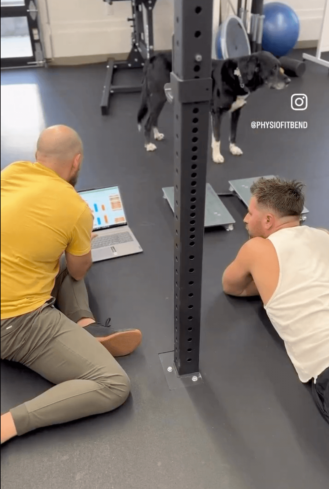 Rear view of a client lying prone on the gym floor, immediately reviewing performance metrics on a laptop in front of a physical therapist sitting beside him. VALD force plates are visible directly in front of the client.