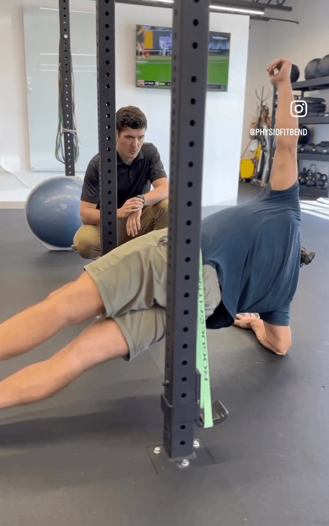A physical therapist watching intently as a client performs a side plank. The view is from behind the client, focusing on the PT's face and coaching expression.